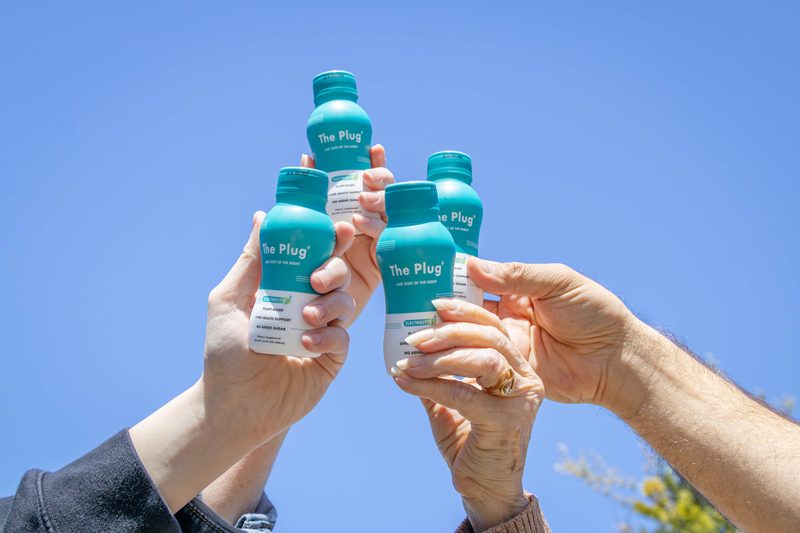 Friends raising bottles of The Plug Drink against a blue sky