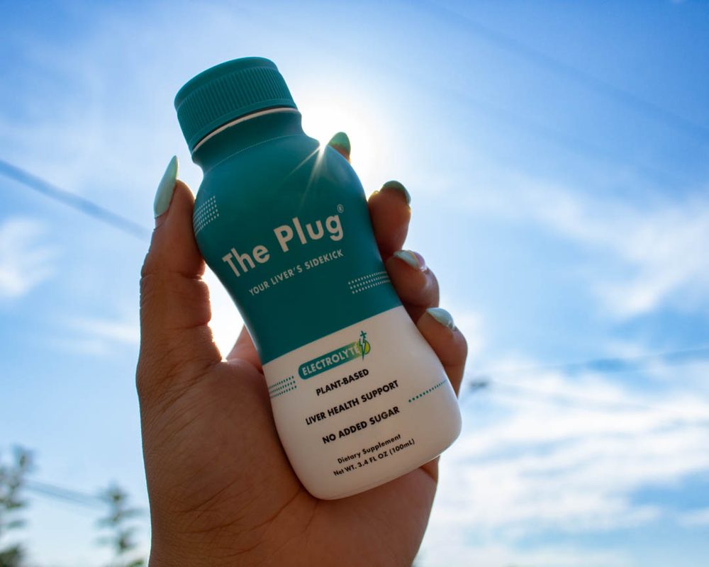 A hand holding a bottle of The Plug Drink against a bright sky